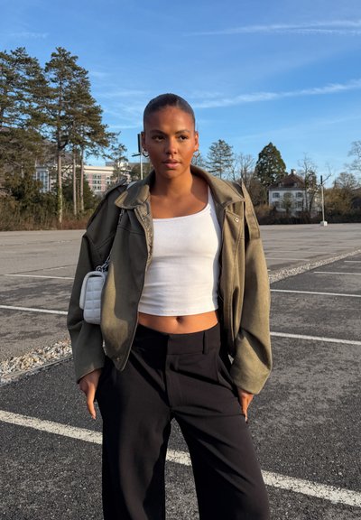 Joven mujer de pie en un estacionamiento vacío, lleva una chaqueta verde, top corto blanco, pantalones negros y un pequeño bolso blanco sobre el hombro, con árboles al fondo.