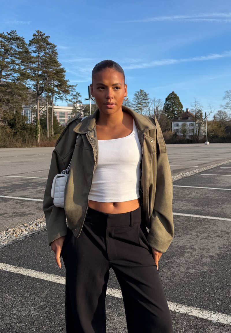Joven mujer de pie en un estacionamiento vacío, lleva una chaqueta verde, top corto blanco, pantalones negros y un pequeño bolso blanco sobre el hombro, con árboles al fondo.