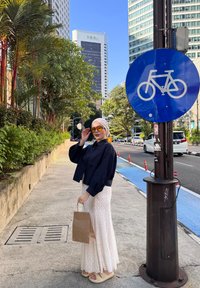 Vrouw in hijab en zonnebril, met een tas, staat op een stoep nabij een blauw fietspadbord en een stadsstraat met hoge gebouwen.