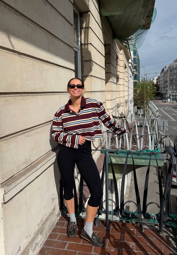 Mujer con gafas de sol y camisa a rayas se apoya en la barandilla del balcón de un edificio beige soleado en una calle urbana.