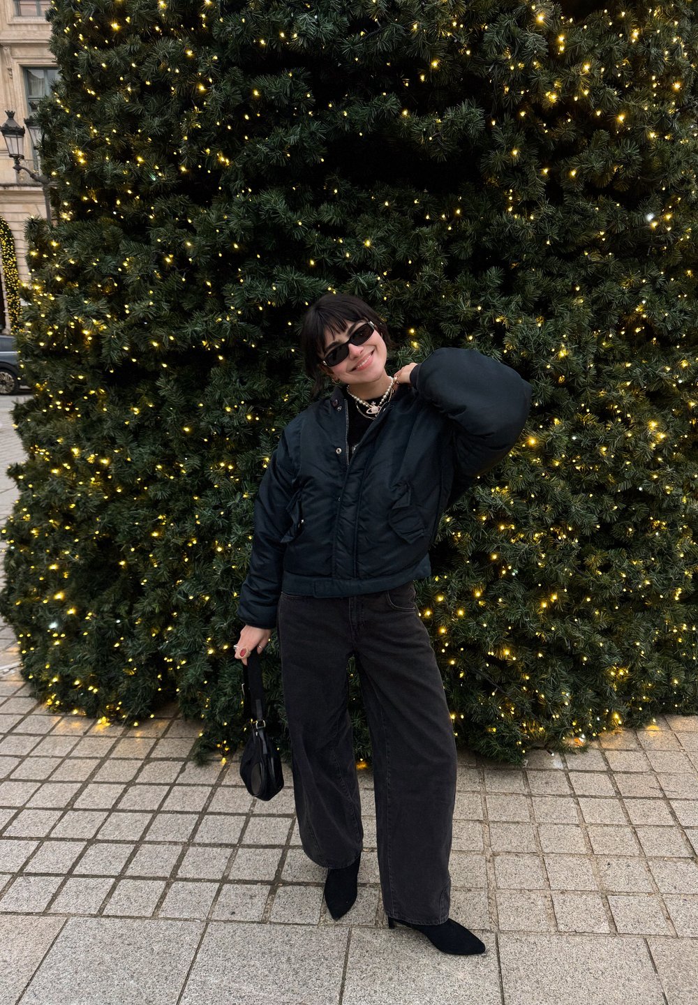 Jeune femme en veste et pantalon noirs posant avec un sac à main devant un grand sapin de Noël décoré de lumières jaunes.