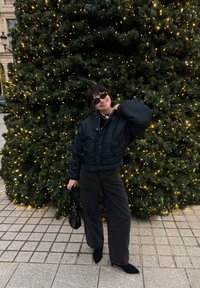 Jeune femme en veste et pantalon noirs posant avec un sac à main devant un grand sapin de Noël décoré de lumières jaunes.
