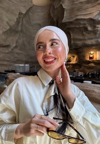 Donna sorridente che indossa un foulard bianco e una camicia tiene degli occhiali da sole, davanti a uno sfondo interno dalla texture simile a una grotta con illuminazione soffusa.