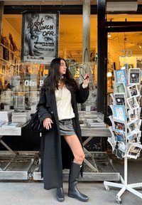 Žena v černém kabátu a botách držící sluneční brýle stojí před knihkupectvím s stojanem na pohlednice a plakátem k uvedení knihy.