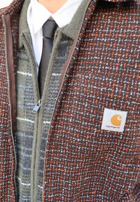 Veste Carhartt à carreaux marron et orange avec doublure en polaire, couche intérieure grise à motif, fermeture éclair argentée et écusson logo brodé.