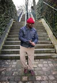 Άνδρας που φοράει κόκκινο σκουφί, γυαλιά, τζιν μπουφάν, μπεζ παντελόνι και καφέ παπούτσια, στέκεται σε πλακόστρωτο μονοπάτι μπροστά από πέτρινα σκαλοπάτια με κουπαστές.