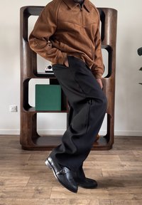 Personne appuyée contre une étagère en bois, portant une veste marron, un pantalon large noir et des chaussures en cuir noir sur un sol en bois.