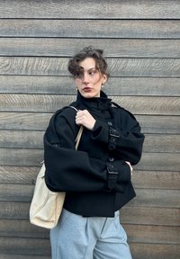 Sort oversized jakke med klokkeærmer, knapper og justerbare spændebånd. Kombineret med grå bukser og en lys beige skuldertaske.