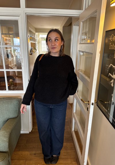 Mujer con suéter negro y jeans de pie en un marco de puerta dentro de una habitación acogedora y bien iluminada con pisos de madera y un sofá verde.