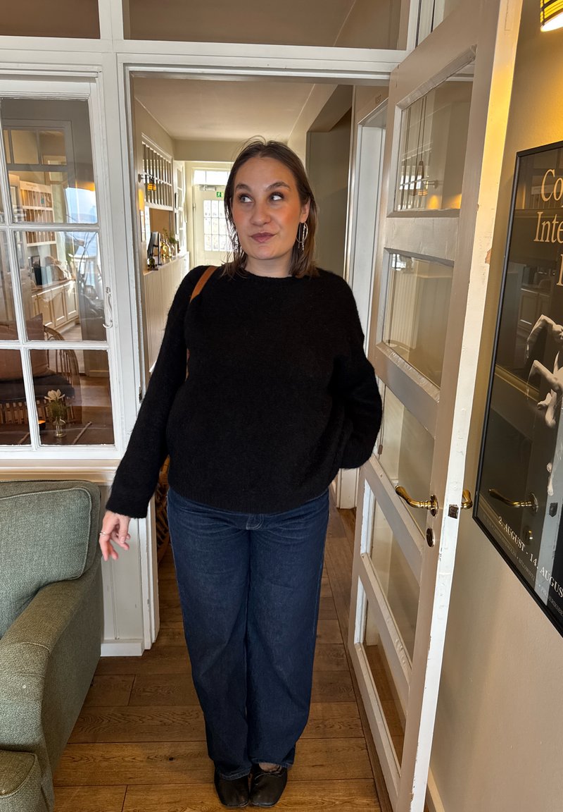 Donna con maglione nero e jeans in piedi in una porta all'interno di una stanza accogliente e ben illuminata con pavimenti in legno e un divano verde.