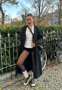 Zwarte overjas over een wit hemd, gecombineerd met pailletten shorts, zwarte maillots en sneakers. Op de achtergrond groenvoorziening en een fiets.