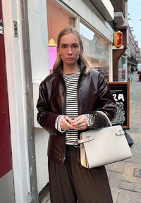Giacca in pelle lucida marrone sopra una maglietta a righe nero e bianco, abbinata a pantaloni larghi neri e una borsa grigia chiaro con dettagli in argento.