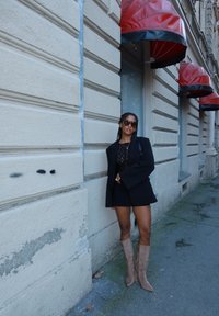 Czarny, oversize'owy blezer, koronkowa bluzka, spódnica oraz beżowe kozaki za kolano. Na ścianie fakturowana farba; nad głową czerwone markizy.