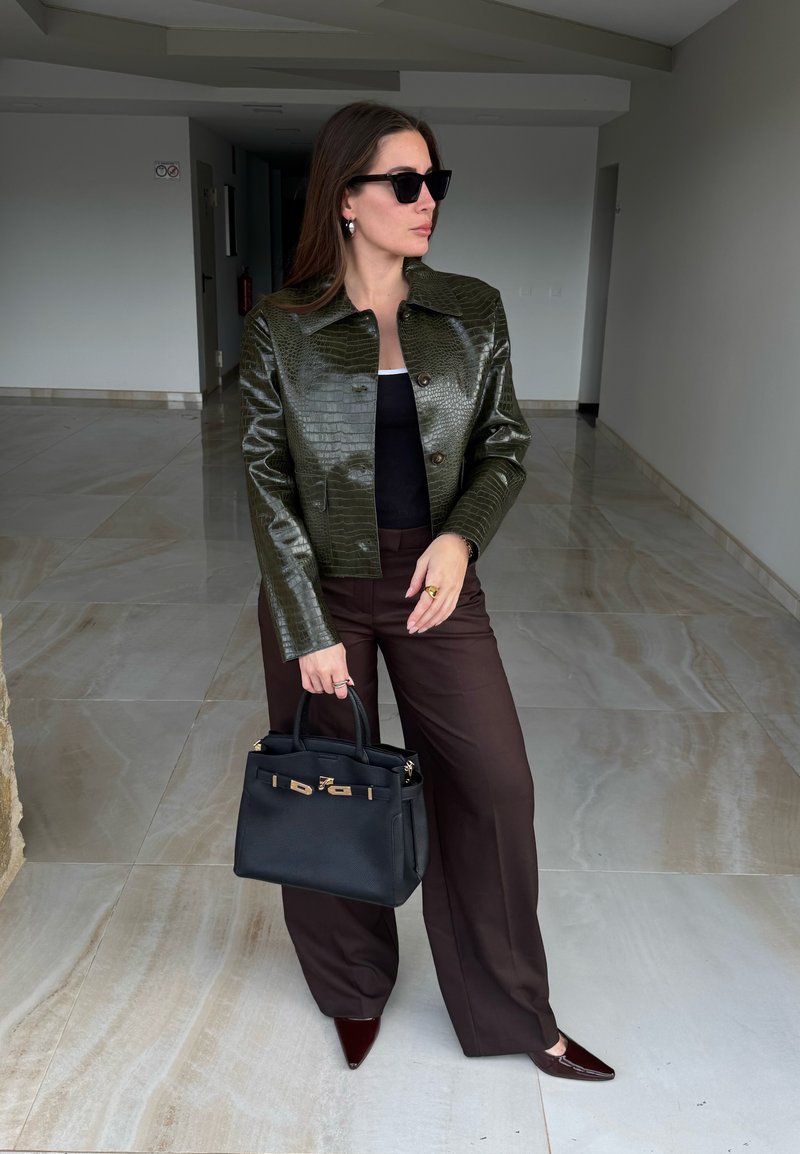 Femme portant une veste en cuir synthétique vert foncé façon crocodile, un haut noir, un pantalon large marron, des chaussures pointues, des lunettes de soleil, et tenant un sac à main noir à l'intérieur.