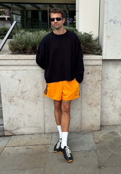 Sudadera negra de gran tamaño, pantalones cortos naranjas con rayas negras, calcetines blancos y zapatillas negras con acentos naranjas. De pie contra una pared de piedra.