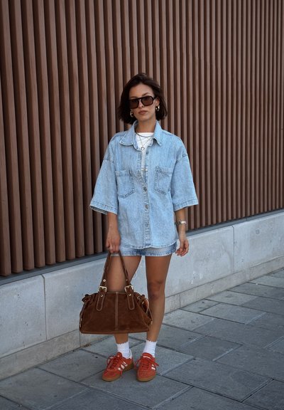 Camisa oversized de mezclilla azul claro, camiseta blanca debajo, bolso de ante marrón, zapatillas rojas, calcetines blancos, de pie contra un fondo de madera.