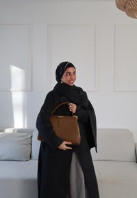 Sac à main en cuir marron à forme structurée avec des garnitures en or, associé à un manteau rayé foncé et une écharpe. Canapé gris doux en arrière-plan.