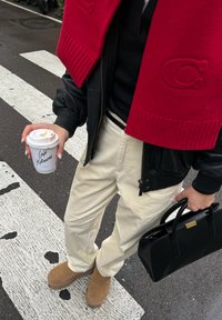 Melna ādas bombējama jaka, sarkans adīts šalle, melna krekls, balti kordurojas bikses, brūnas kurpes; tur kafijas krūzi un melnu rokassomiņu.