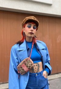 Casaco trench azul, blusa justa azul e calças em veludo cotelê. Chapéu castanho. Bolsa transversal multicolorida com padrão de logótipos. Cabelo vermelho com detalhes pretos.