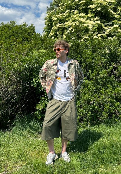 Chaqueta de camuflaje floral, camiseta blanca con estampado gráfico, pantalones cortos oversize verde oliva, zapatillas grises y gafas de sol naranjas. Fondo de follaje verde.