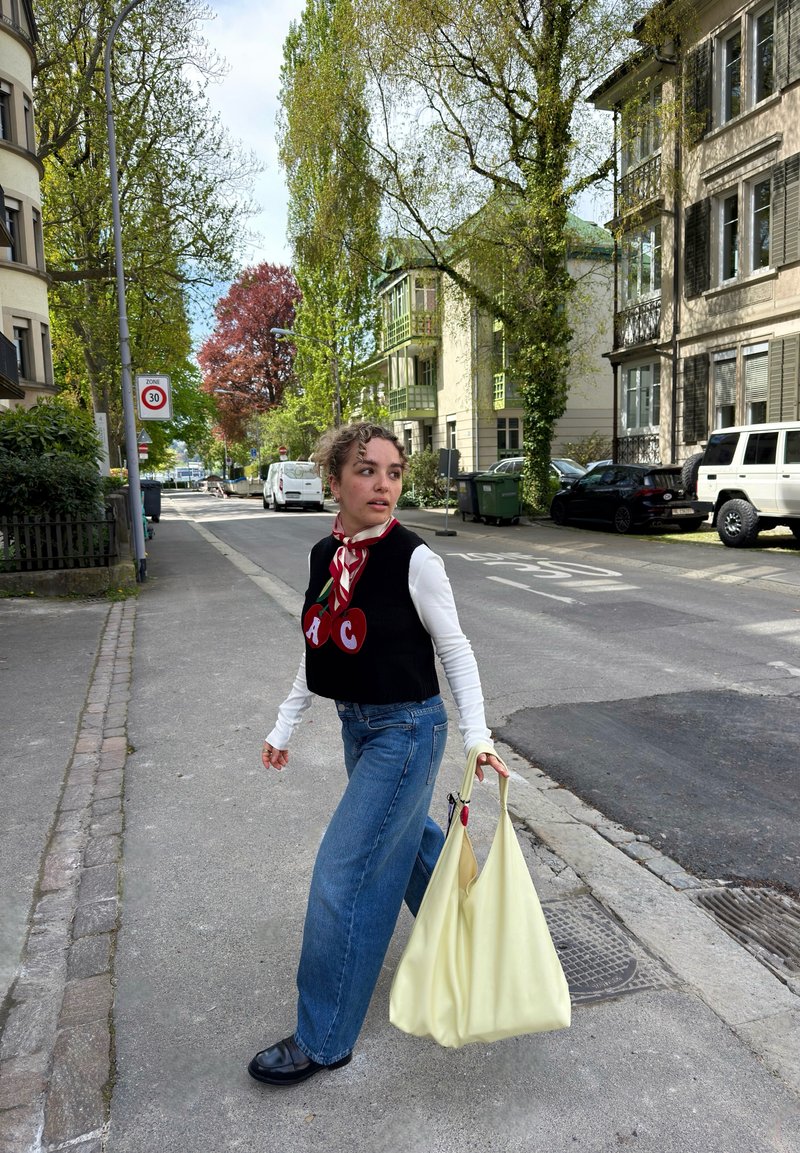 Ung kvinde krydser en stille boligvej, iført en sort vest med kirsebær, blå jeans, sorte sko og bærer en stor lysegul taske.