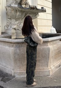 Black handbag with metallic hardware, beige fluffy jumper, camouflaged trousers, standing by a stone fountain with water flowing.