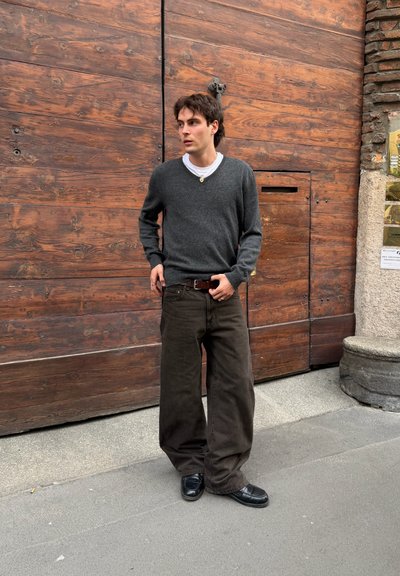 Hombre con suéter gris oscuro, pantalones marrones holgados y zapatos negros, de pie en la acera frente a una gran puerta de madera.