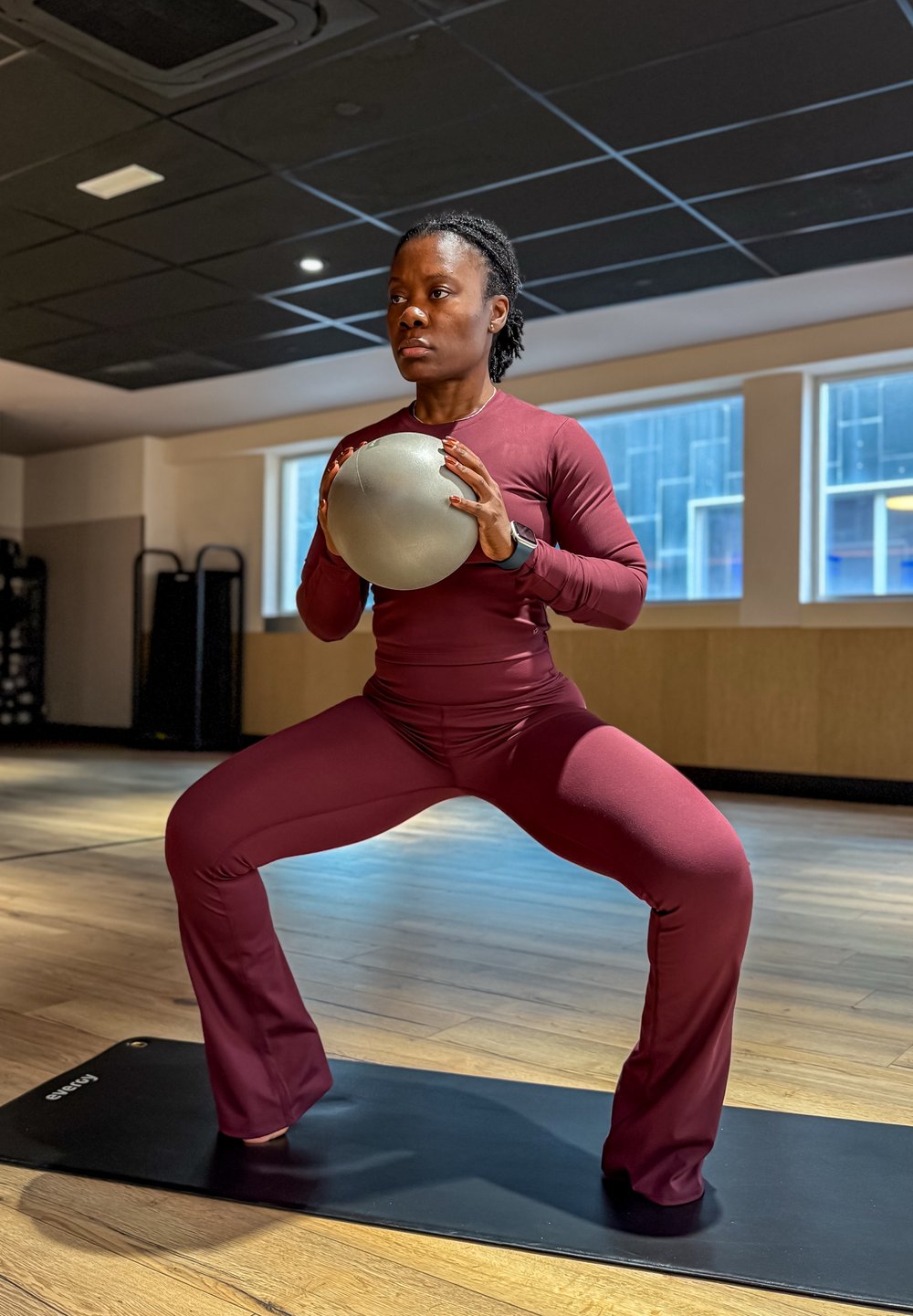 Ensemble d’entraînement bordeaux, comprenant un haut à manches longues et un pantalon ajusté, avec un ballon d’exercice gris texturé, posé sur un tapis de yoga noir dans une salle de sport.