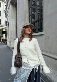 Pull blanc en maille avec boutons, associé à une superposition de dentelle et de denim. Une casquette marron clair et un sac à bandoulière marron complètent le look.