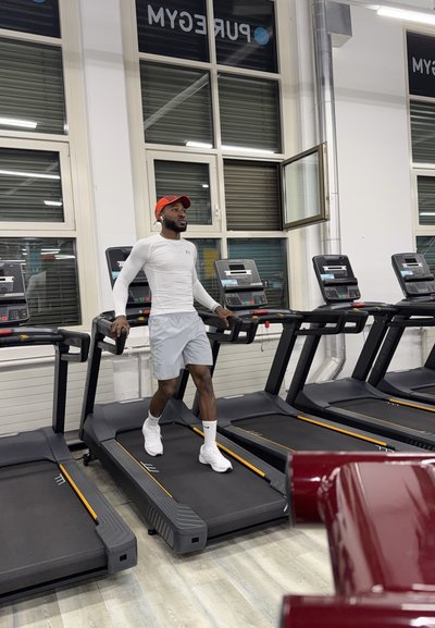 Hombre caminando en una cinta de correr en el gimnasio, vistiendo una camiseta blanca de manga larga, pantalones cortos grises, zapatillas blancas y gorra roja con auriculares.