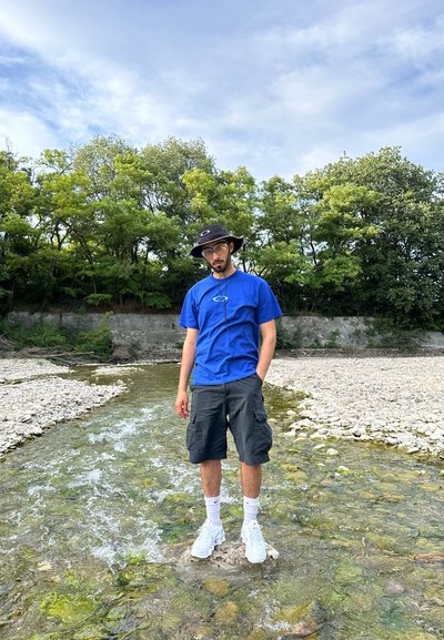 Camiseta azul, pantalones cortos de cargo negros, zapatillas blancas y un sombrero negro de ala ancha. De pie sobre una roca en un arroyo poco profundo con árboles verdes al fondo.