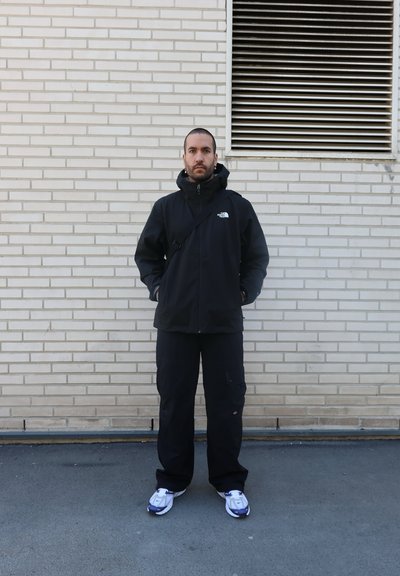 Chaqueta negra impermeable con capucha, frente con cremallera y logo. Pantalones cargo negros y zapatillas blancas con acentos morados. De pie frente a una pared de ladrillo.