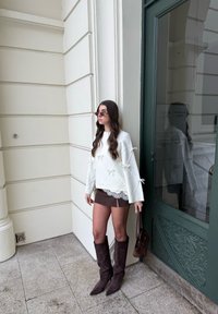 Femme aux longs cheveux bruns portant un pull blanc avec un nœud, une mini-jupe brune, de hautes bottes brunes et des lunettes de soleil, debout près d'une porte verte en extérieur.