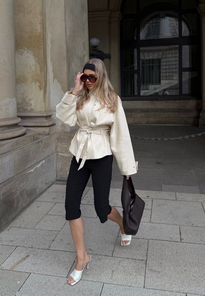 Mujer con chaqueta beige con cinturón, pantalones cortos negros, sandalias blancas de tacón, gafas de sol grandes, sosteniendo un bolso marrón oscuro caminando sobre pavimento de piedra.