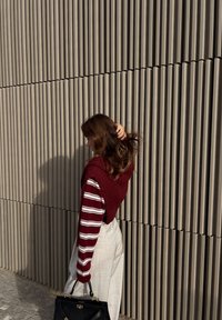 Pulover roșu din tricot cu model, purtat peste un top cu mâneci lungi în dungi, asortat cu pantaloni evazați de culoare deschisă, ținând o geantă neagră, în fața unui perete texturat.