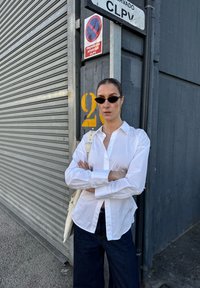 Biała koszula z guzikami, w oversize'owym fasonie, noszona z ciemnymi szerokimi spodniami. Uzupełniona czarnymi okularami przeciwsłonecznymi i kremową torebką.