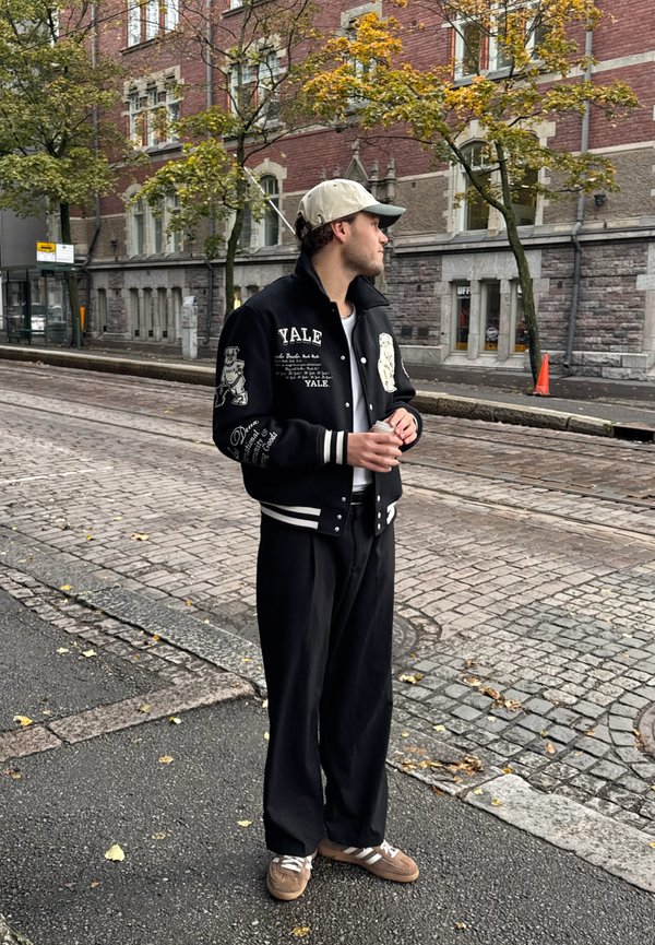 Veste universitaire noire avec des rayures blanches, ornée de logos et de broderies de Yale ; associée à un pantalon noir ample et des baskets marron. Casquette avec une visière verte.