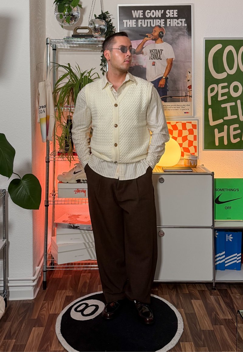 Kremasti pleteni prsluk preko prugaste košulje, smeđe krojene hlače i crne cipele. U pozadini se nalaze biljke i plakati, uz toplo osvjetljenje.