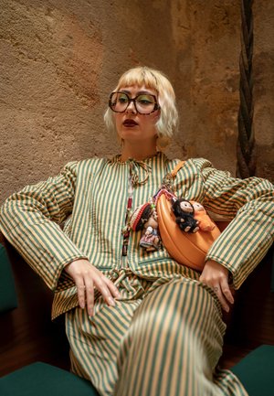 Conjunto de dos piezas a rayas en verde y amarillo, con un bolso naranja redondeado adornado con pequeñas figuras colgantes. Gafas grandes y estampadas, y cabello corto.