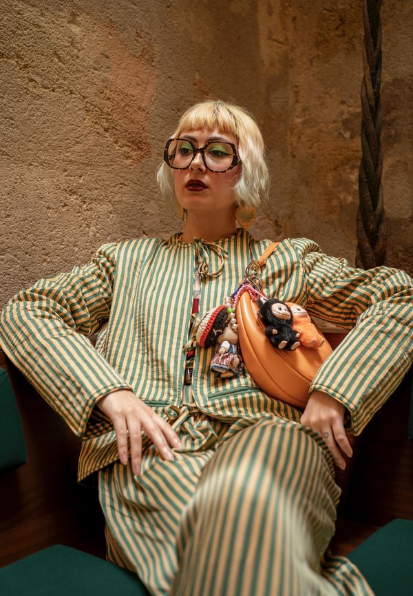 Ensemble deux pièces rayé vert et jaune avec un sac orange arrondi orné de petites figurines suspendues. Grandes lunettes à motif et cheveux courts.