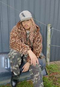 Plašč iz umetnega krzna z leopardjim vzorcem, kombiniran s hlačami s kamuflažnim vzorcem. Dodatki vključujejo sive kape in očala, postavljene na sivi steni.