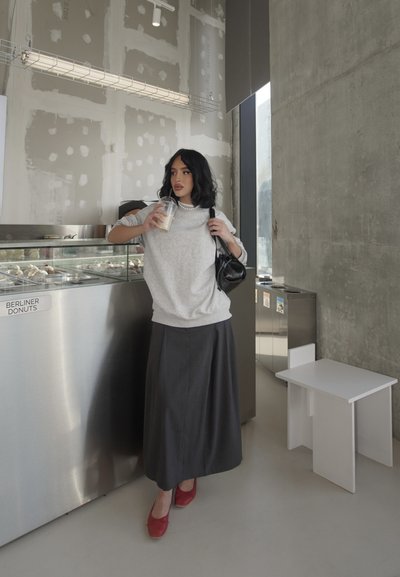 Sudadera gris, falda larga oscura, zapatos rojos; sosteniendo una bebida helada. Café minimalista con mostrador de acero inoxidable y pared sin terminar.
