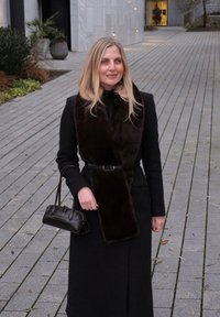 Long manteau noir avec un col en fourrure foncée, taille cintrée avec une ceinture, sac à main noir à finition texturée, debout sur un chemin pavé.