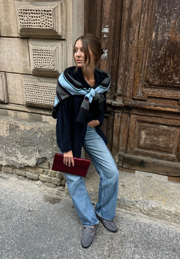 Une femme en manteau sombre, avec une écharpe rayée et un jean à jambes larges, tient une pochette rouge à côté d’une porte en bois patinée.