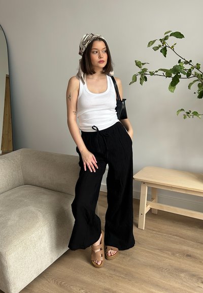 Pantalones anchos plisados negros, top blanco sin mangas, sandalias marrones. Modelo con cabello corto y pañuelo de cabeza con estampado, de pie junto a una mesa de madera clara.