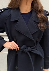 Marinblå dobbelspent ullfrakk med store knapper, belte i midjen og skulderklaffer. Myk tekstur og skreddersydd design.