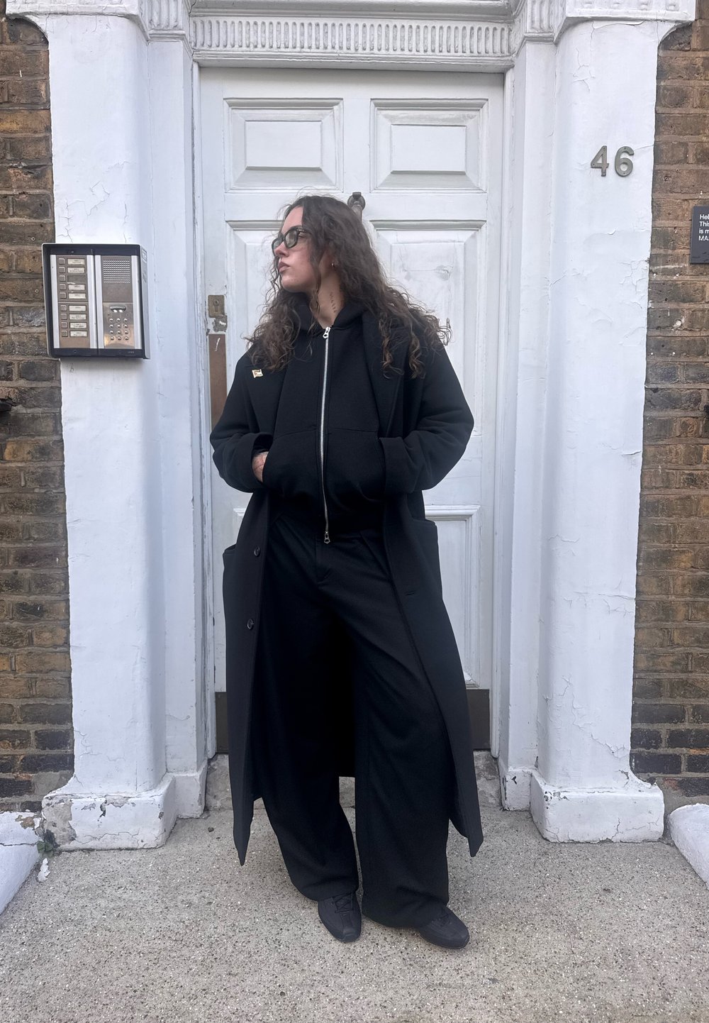 Long manteau noir porté sur un sweat à capuche noir à fermeture éclair et un pantalon large, assortis à des chaussures noires. Mur en pierre et porte blanche en arrière-plan.