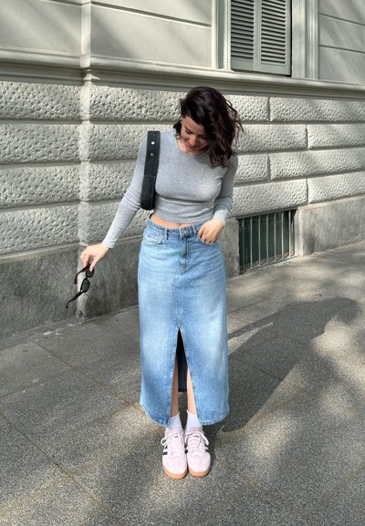 Top de manga larga gris claro, falda midi de mezclilla con abertura frontal y zapatillas rosa pálido con detalles blancos. Gafas de sol en la mano.
