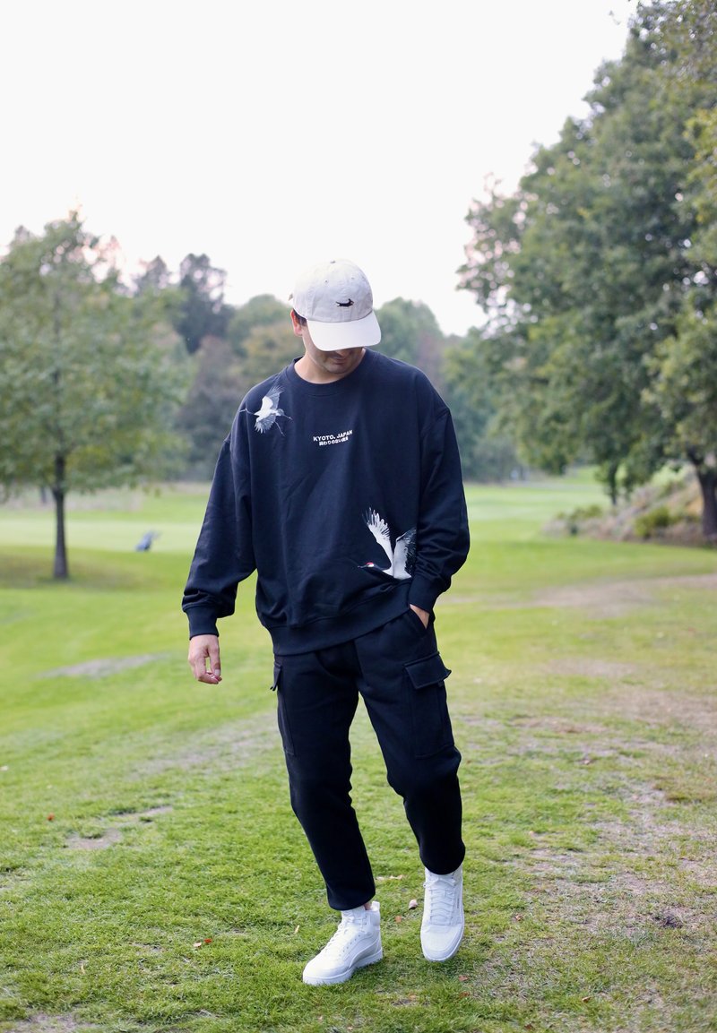 Svart sweatshirt med vita kran-tryck, kombinerat med svarta cargobyxor och vita sneakers. Justerbar vit keps som bärs bak och fram.