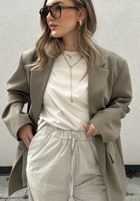 Blazer beige oversize, t-shirt bianco e pantaloni rigati beige chiaro. Gli accessori includono occhiali da sole grandi e collane d'oro a strati.